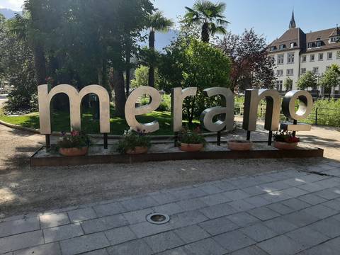 Large metal letters spelling 'merano' set in a leafy town park with colorful flowers.