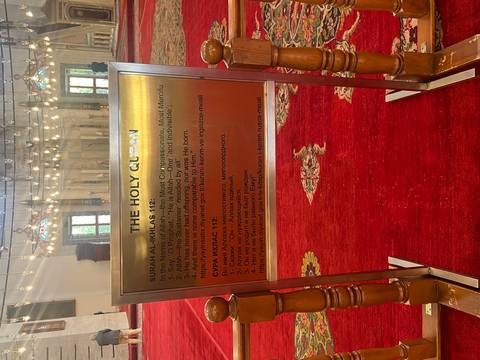 Multilingual sign explaining a Quran chapter stands on a red-carpeted mosque floor.