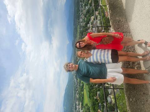 Three friends smile at a panoramic viewpoint overlooking a valley of alpine villages and peaks.