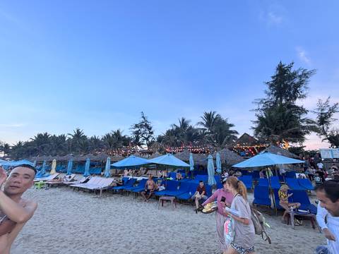A lively beach scene at dusk with rows of blue loungers, thatched umbrellas, and holidaymakers enjoying the shoreline.