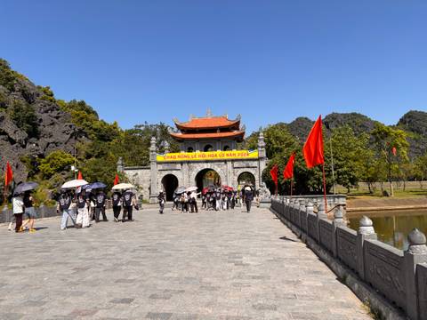 Schoolchildren walk with umbrellas toward a historic stone gate flanked by red flags at Hoa Lu complex.