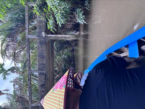 A traveller wearing a conical hat paddles a blue sampan beneath a low concrete bridge through muddy jungle waters.