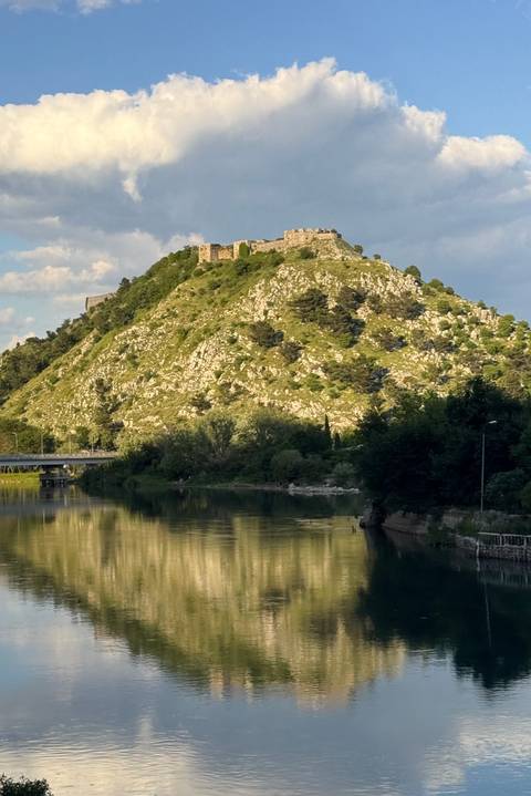 A green, rocky hill topped with a stone fortress rises above a calm river and bridge at golden hour.