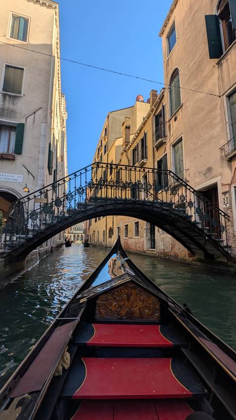 View from gondola prow of ornate iron footbridge arching over narrow Venetian canal