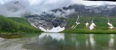 Serene alpine lake with melting snow patches beneath misty peaks reflected in green water.