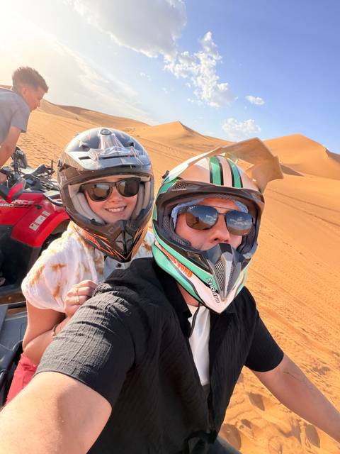 Selfie of two people wearing helmets on quad bike against orange desert dunes