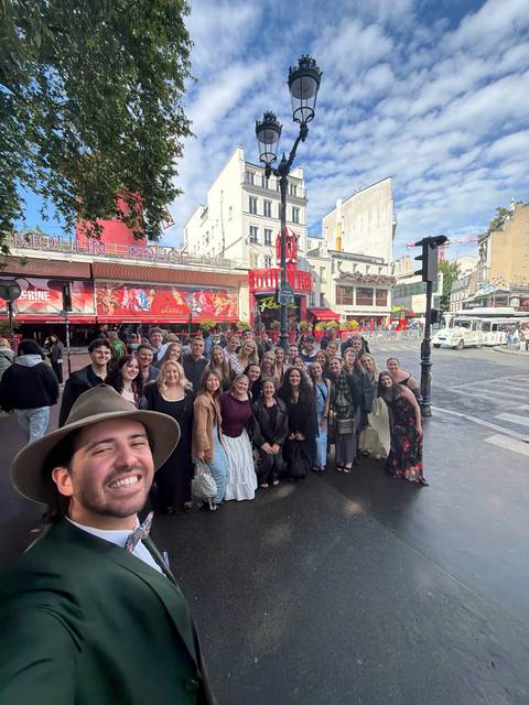 Large tour group posing and smiling in front of the iconic red windmill façade of the Moulin Rouge cabaret on a sunny day in Paris, with a man taking a selfie in the foreground.