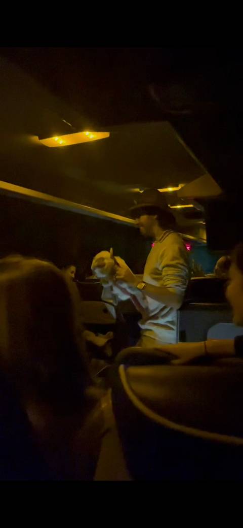 A man standing in a dimly lit tour bus aisle playfully holding a small stuffed animal while fellow passengers sit around him.