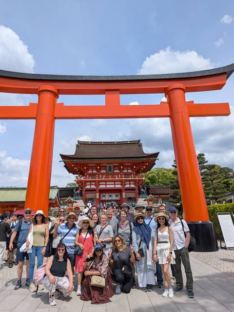 Tour group gathered beneath a massive vermilion torii gate with the ornate entrance of Fushimi Inari Shrine behind them in Kyoto.