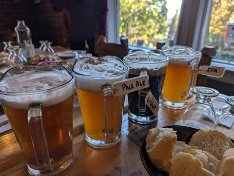 Flight of craft beers in labelled glass mugs arranged on a wooden table in a rustic restaurant.