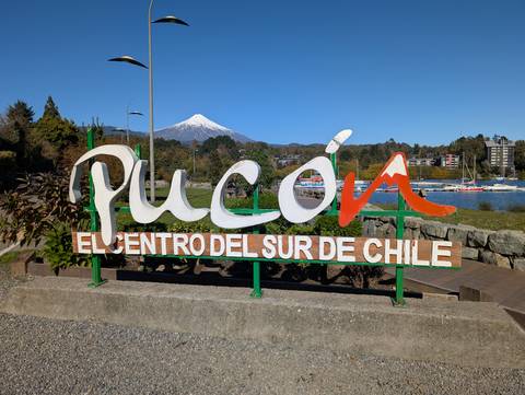 White-and-orange 'Pucón' sign stands in front of a lake and snow-capped volcano under clear skies in southern Chile.