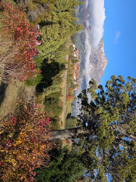 Colourful autumn shrubs, green fairways and tall trees frame a lodge complex beneath a rugged mountain peak against a bright blue sky.