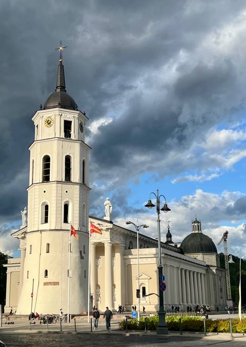 Tall white bell tower with statues and national flags under a dramatic cloud-filled sky.