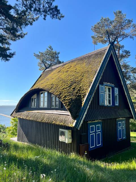 Traditional wooden cottage with steep moss-covered roof overlooking the Curonian Lagoon on a sunny day.