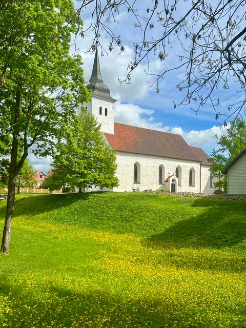 A small white church with a red-tiled roof surrounded by leafy green trees on a sunny day