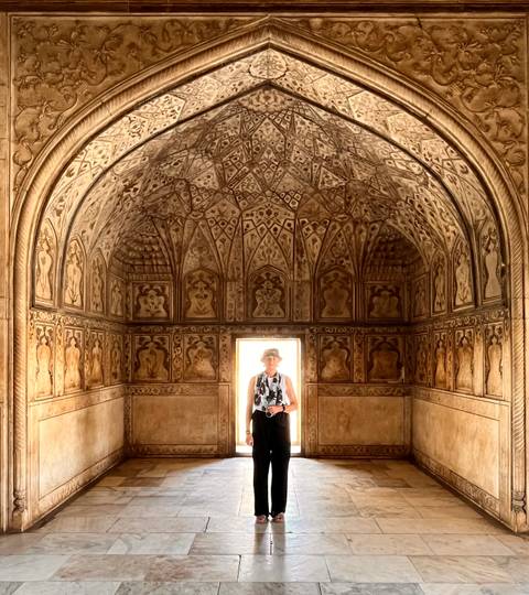 An older traveler stands in a richly decorated Mughal chamber with intricate stucco patterns and vaulted ceiling.