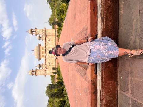 Traveler poses on red sandstone terrace with Itimad-ud-Daulah mausoleum (Baby Taj) in the background under blue sky.