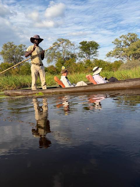 Guide poling a mokoro while two tourists recline on cushions, reflected in calm Okavango Delta waters.