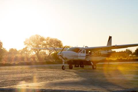 Light aircraft parked on sandy runway at sunrise with warm lens flare and silhouettes of trees.