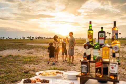 Family enjoying sundowner drinks in the savannah at sunset with liquor bottles in foreground.