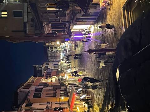 A lively Venetian street at night is lit by shopfronts and cafés as people stroll along wet cobblestones.