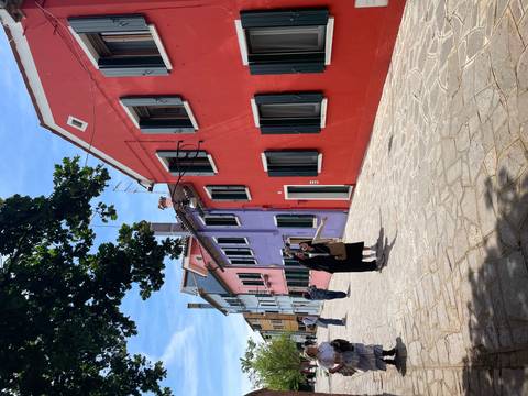 Colorful red, purple and blue houses with dark shutters line a sunny lane on Burano island.