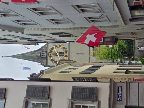 Narrow Zurich lane framed by tall buildings leading to the large clock face of St. Peter’s church with Swiss flag