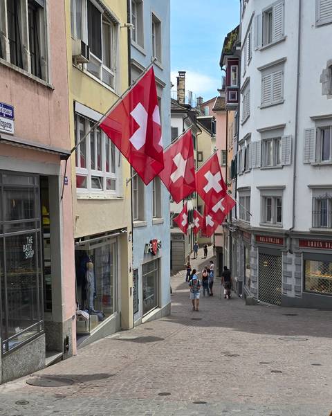 Row of Swiss flags hanging over a narrow pedestrian street with small shops and strolling visitors