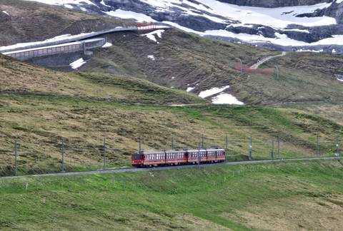 Red cog railway train climbing green alpine slopes with patches of late-season snow