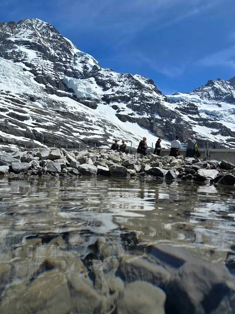 Low-angle view across rocky melt-water pool toward tourists on a platform beneath snowy peaks