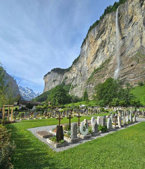 Alpine cemetery set beneath towering cliff and thin waterfall with snow-tipped peaks beyond