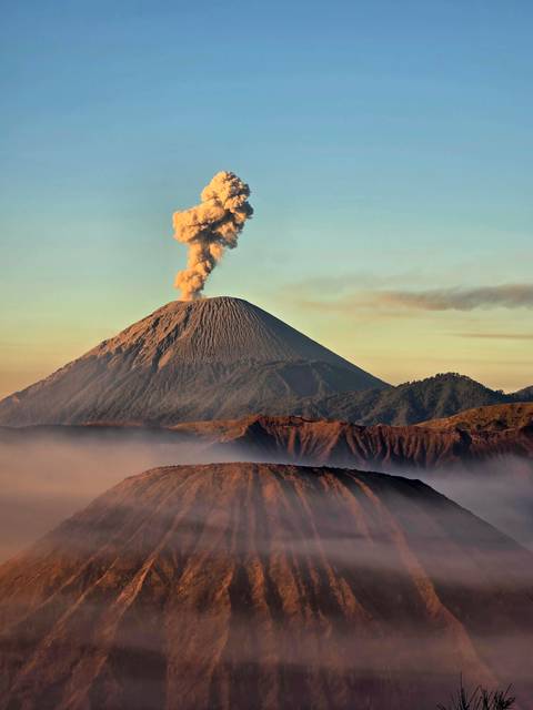 Active volcano emitting thick ash plume above ridged cone at sunrise with misty caldera below