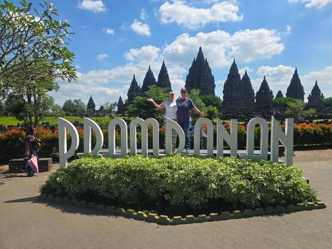 Travellers posing atop landscaped sign spelling Prambanan with temple spires in background