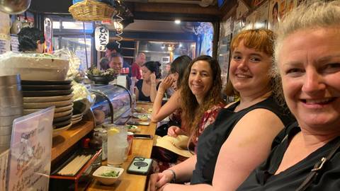 Lively diners seated shoulder-to-shoulder at a small Japanese izakaya counter filled with dishes and lanterns