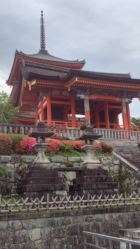 Vibrant vermilion temple gate framed by stone lanterns and blooming shrubs on a cloudy day in Kyoto