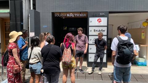 Guide speaks to a cluster of travelers outside a modern building labeled Solasia Bldg Tsukiji in Tokyo