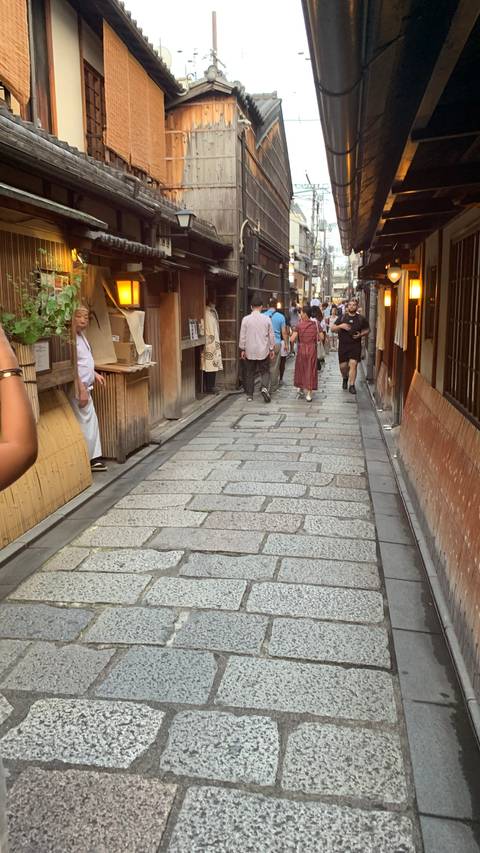 Stone-paved alleyway flanked by wooden facades where pedestrians stroll past intimate restaurants at dusk
