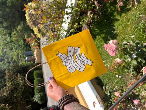 Close-up of a yellow Fragonard shopping bag held outdoors near blooming flowers.