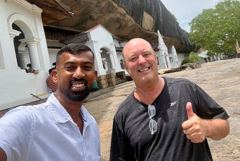 Two men smiling for selfie at cave-temple complex built under giant rock overhang.