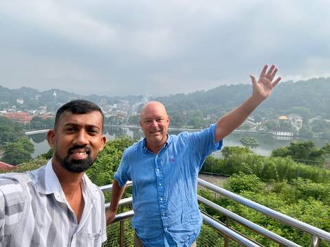 Two men taking selfie on a balcony overlooking misty Kandy lake and hills.