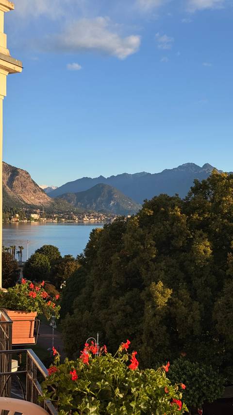Serene lake bordered by wooded hills and distant alpine peaks under clear blue sky.