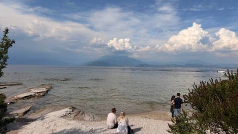 Small group relaxing by a rocky lakeshore with billowing clouds over distant peaks.