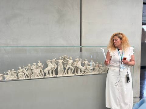 Tour guide explains a classical frieze replica inside a museum, holding a microphone and notes
