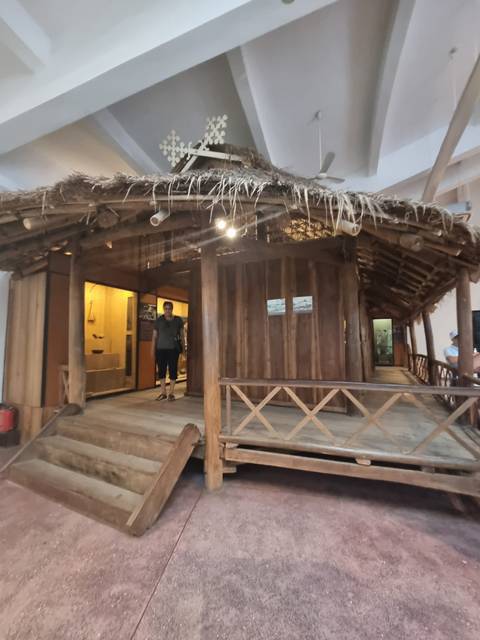 Visitor standing inside a rustic wooden stilt house exhibit with bamboo roof and wooden railings.