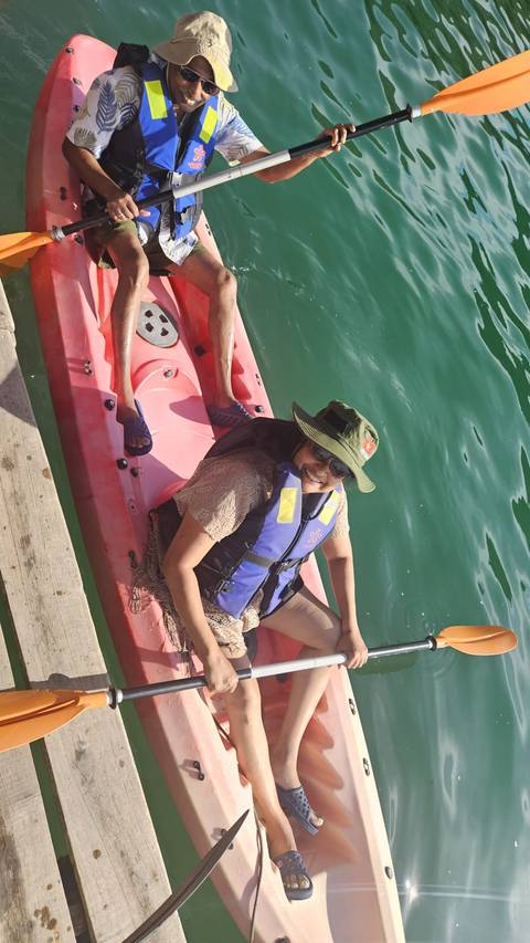 Two travellers in a red kayak on emerald water, one smiling toward the camera wearing a life jacket and sun hat.