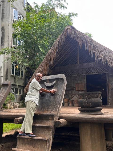 Smiling man posing with a large carved wooden gong outside a traditional thatched hut surrounded by pottery.