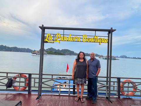 Couple posing on a pier beneath a 'La Pandora Boutique' sign with Halong Bay's karst islands and cruise boats behind.
