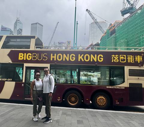 Couple posing beside a maroon double-decker Big Bus Hong Kong tour bus on a cloudy day.