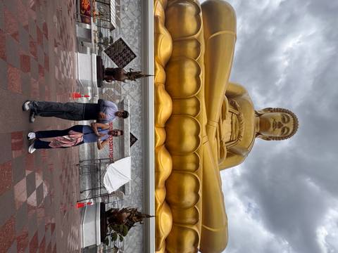 Travellers stand in prayer pose beneath a towering golden seated Buddha statue under dramatic grey clouds.