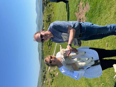 Smiling couple gently hold and feed a lamb on a sunny green hillside farm in Ireland.